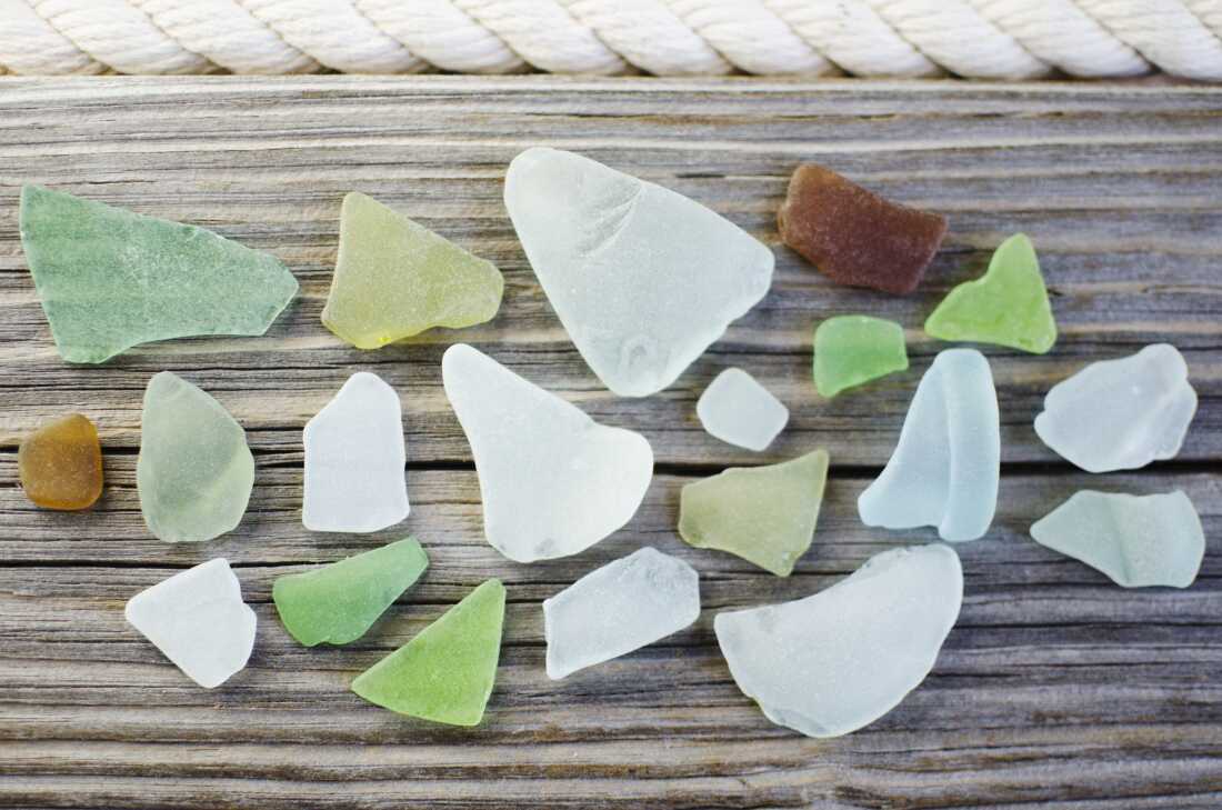 A close-up, high-angle photograph showing various colors of green, blue, white, and brown sea glass neatly arranged on a wooden surface.  