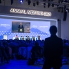 In this photo, President Trump appears on large screens while giving an address via video conference. In a large room, attendees of the World Economic Forum's annual meeting sit in rows of chairs, watching the screens, and five people sit on a stage.