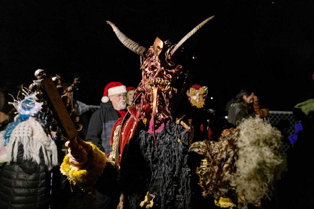 A man dressed in a large, horned mask with a long tongue holding a large a long stick with bells on the end.