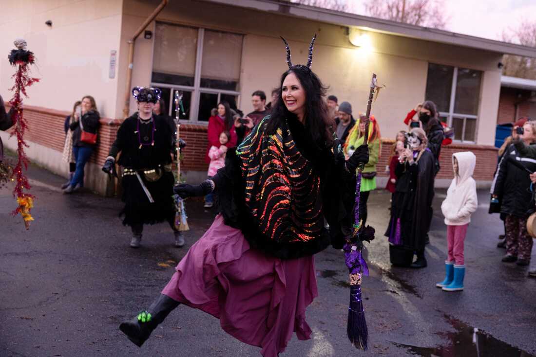 A woman in a long purple skirt and brightly colored shawl dances while wearing horns and holding a broom.