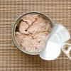 This photo, taken from above, shows a can of tuna. The lid, which has a pull tab, is peeled back, revealing the tuna in the can. The can sits on a rattan-like mat.