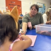 A little girl with her back to the camera sits and draws at a low table the color of blueberries in a classroom. Her preschool teacher leans in with a crayon to help. A plastic bin containing crayons sits between them.