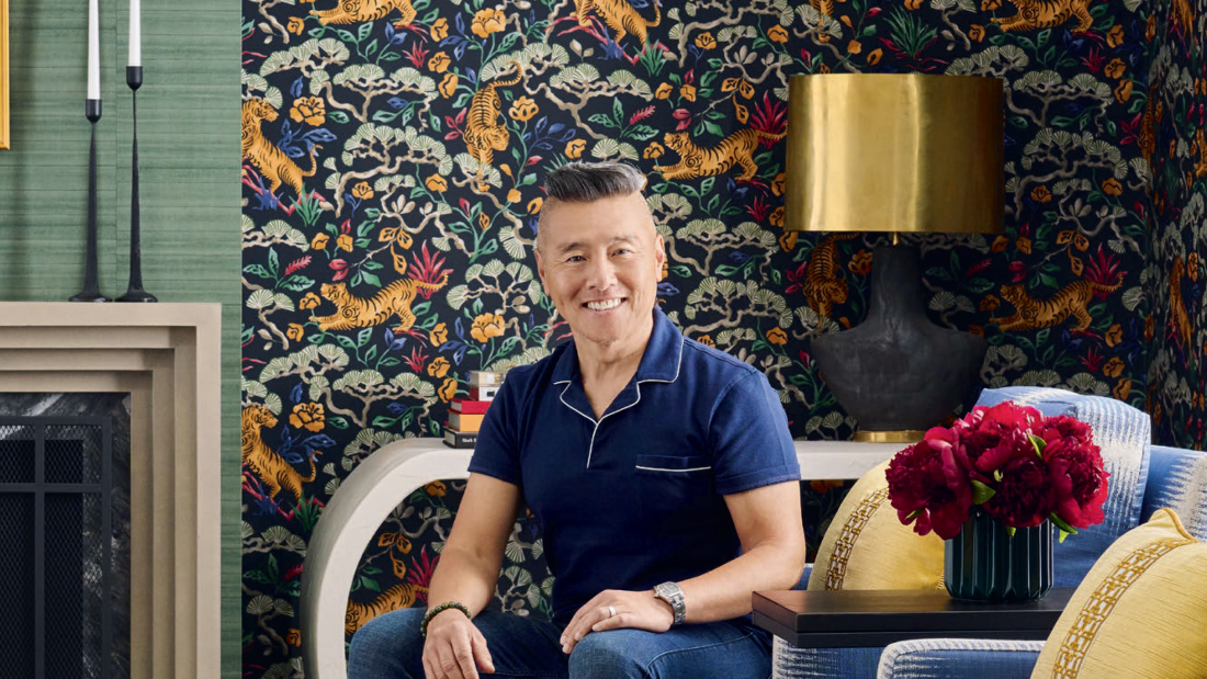 An environmental portrait of Vern Yip sitting on a blue and white patterned couch an aesthetically-pleasing living room he decorated, with tiger and floral patterned wallpaper behind him.