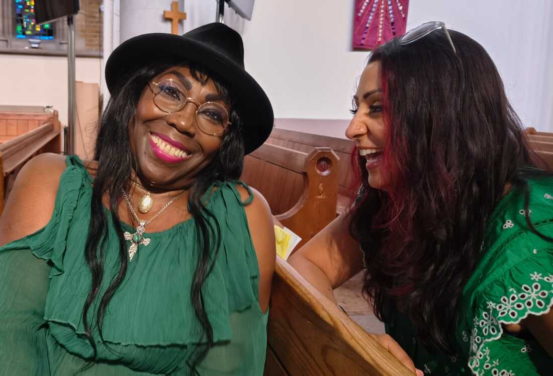 Geraldine Jeffrey (on the left side of the frame) and consultant nurse Sonia Sandhu are shown in a close-up photo. They appear to be in a conversation and are both laughing. They are sitting in church pews, and both are wearing green tops.