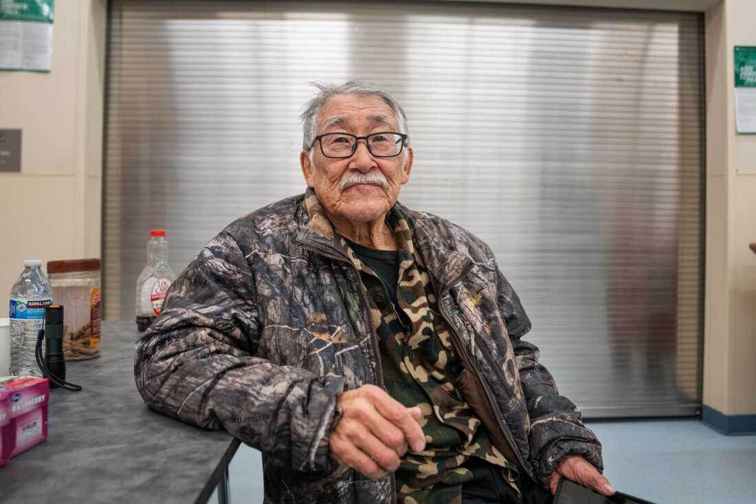 Noah Andrew Sr., 74, sits at a table at the Kwigillingok School. He's wearing a camouflage-patterned shirt and a jacket patterned with leaves and trees.