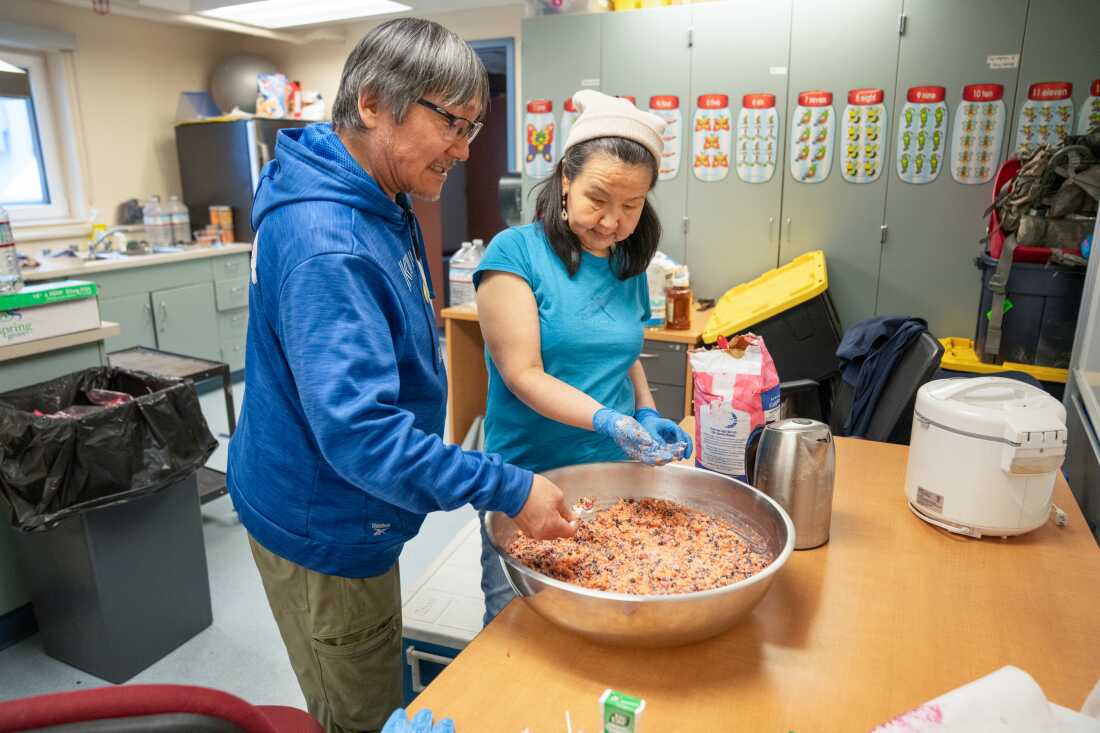 Darrell John stands next to Nettie Igkurak in a kitchen at the Kwigillingok School. They are facing a table on which sits a large metal bowl of Alaskan ice cream, which contains lots of small spherical berries.