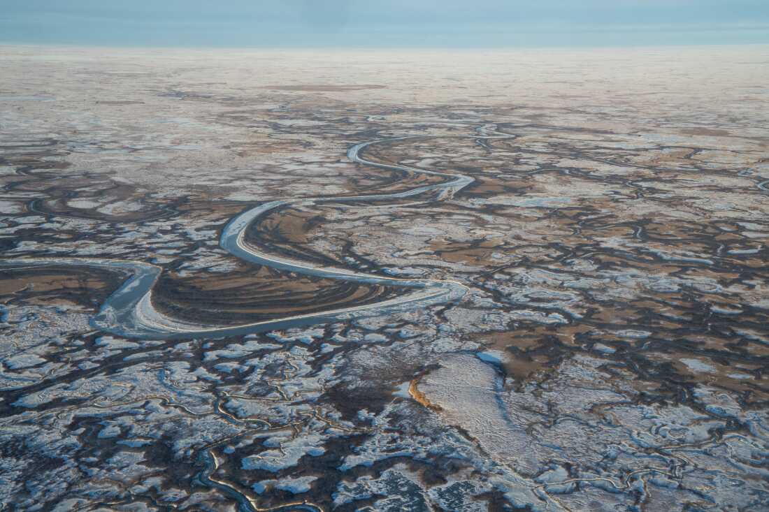 This aerial view shows a vast flat expanse of the Yukon-Kuskokwim Delta in Alaska in October. A body of frozen water snakes through the land, much of which is covered by snow.