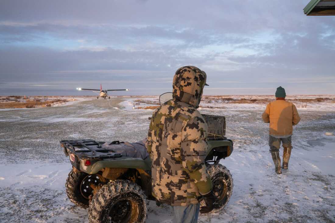 Two men in winter coats stand near an all-terrain vehicle on flat land that's partially covered by snow. A small airplane is on the ground in the background.