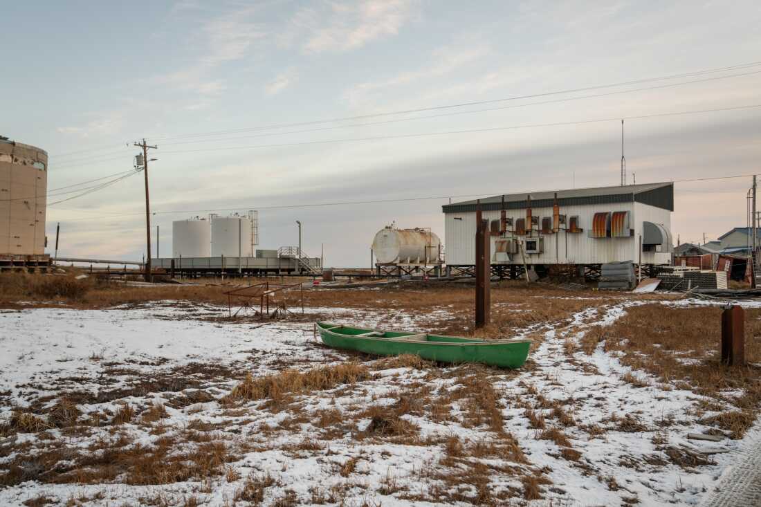 A lone green canoe rests on the land. In the background are three cylindrical storage tanks, one lying horizontal and two standing vertical.