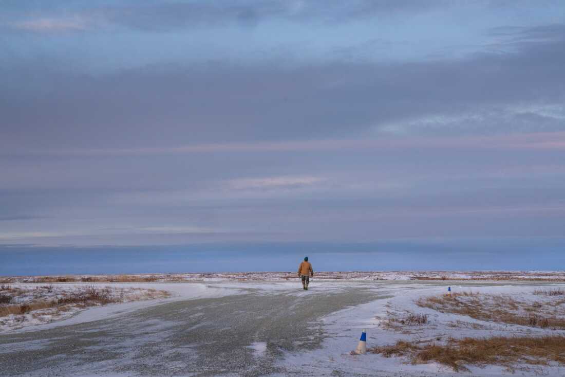A Kwigillingok resident wearing a winter coat walks in the distance, with their back facing the camera. The land is partially covered by snow, and the blue sky has some pink streaks.