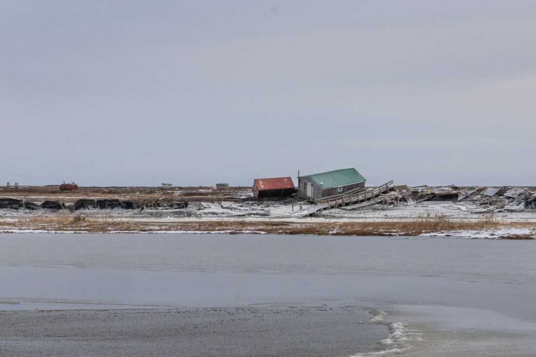 In this photo's foreground is the Kwigillingok River, and on the other side of the river, in the background, are two homes that are tilted askew. Near the homes are a damaged boardwalk and wooden debris. Much of the ground is covered by snow.