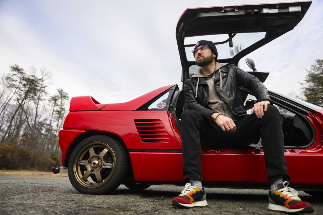Andrew Maxon sits sideways in the right seat of his red Autozam AZ-1 kei car. The gull-wing door is raised upward, and Maxon's feet rest on the ground outside.