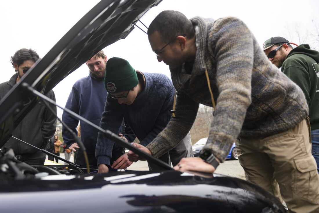 Five car enthusiasts stand near the open hood of a black Suzuki Cappuccino kei car as they work on a minor repair in the engine bay.