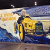 This photo, taken at Berkshire Hathaway's annual shareholder meeting, shows a large display panel that advertises Marmon, a company owned by Berkshire Hathaway. The panel shows a cartoon illustration of Warren Buffett driving an old-fashioned yellow race car on a racetrack.