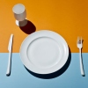 This image shows a high-angle view of a white plate, a stemmed drinking glass, a fork to the plate's right and a knife to the plate's left. The items are all set on a tabletop that's orange across the top of the frame and light blue across the bottom.