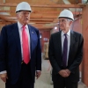 This photo shows President Trump and Federal Reserve Chair Jerome Powell standing next to one another in a large room that's undergoing renovations, with wooden ceiling beams exposed and plywood walls. Both men are wearing white hard hats and suits.