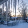 Icicles hang down from the eaves of the roof in the foreground of the photo, looking out onto a snowy neighborhood landscape, including a backyard and surrounding houses, during a winter storm. 
