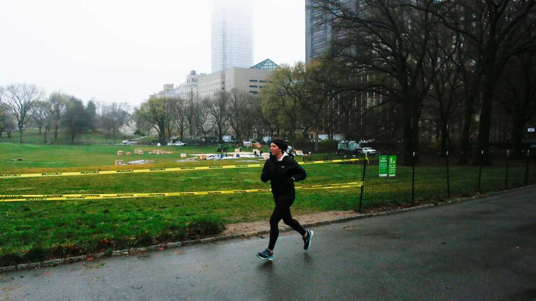 A jogger is seen in warm winter running clothes on a paved path in front of a wide open, green lawn. Buildings are in the background.