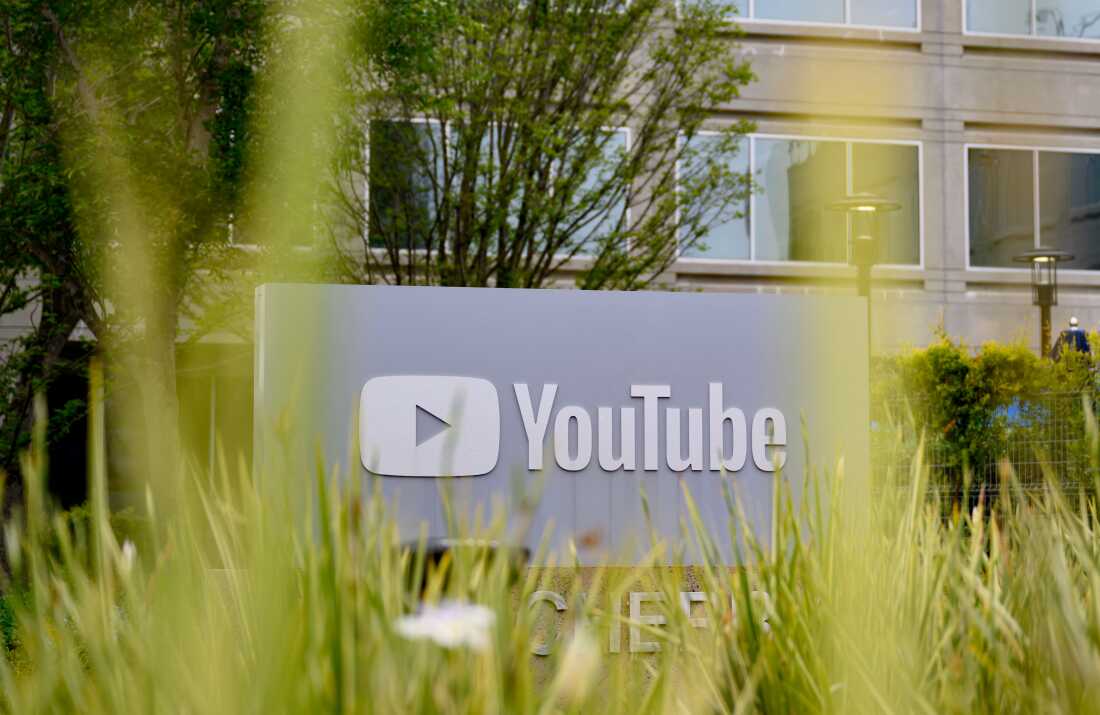 The YouTube logo, featuring a play button, is displayed on a sign outside the company's headquarters in 2025. Blades of grass rise up in the foreground.