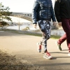 Two people wearing sneakers and photographed from the neck down walk on a paved path that runs along a body of water. A bridge is in the background.