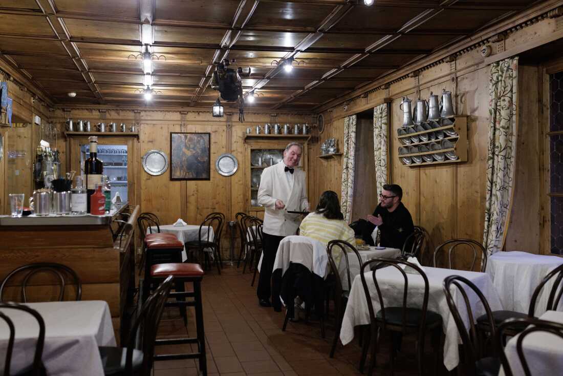 A waiter in a formal white jacket serves patrons inside the wood-paneled Bar Posta at the historic Hotel de la Poste. A cornerstone of Ampezzo social life since the 19th century and a favorite haunt of Ernest Hemingway, the bar represents the "old-world" heritage of Cortina