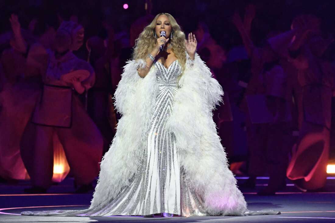 Mariah Carey sings during the opening ceremony. She is wearing a white and silver sparkly gown with white feathers.