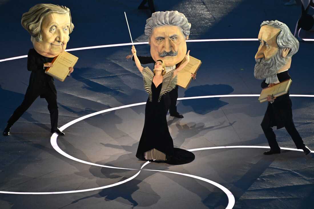 Italian actress Matilda De Angelis (center) performs as a conductor, dressed in a black gown and holding a large baton, with dancers dressed as the three great masters of Italian opera Giuseppe Verdi, Giacomo Puccini and Gioachino Rossini, who are wearing giant heads and holding books of sheet music.