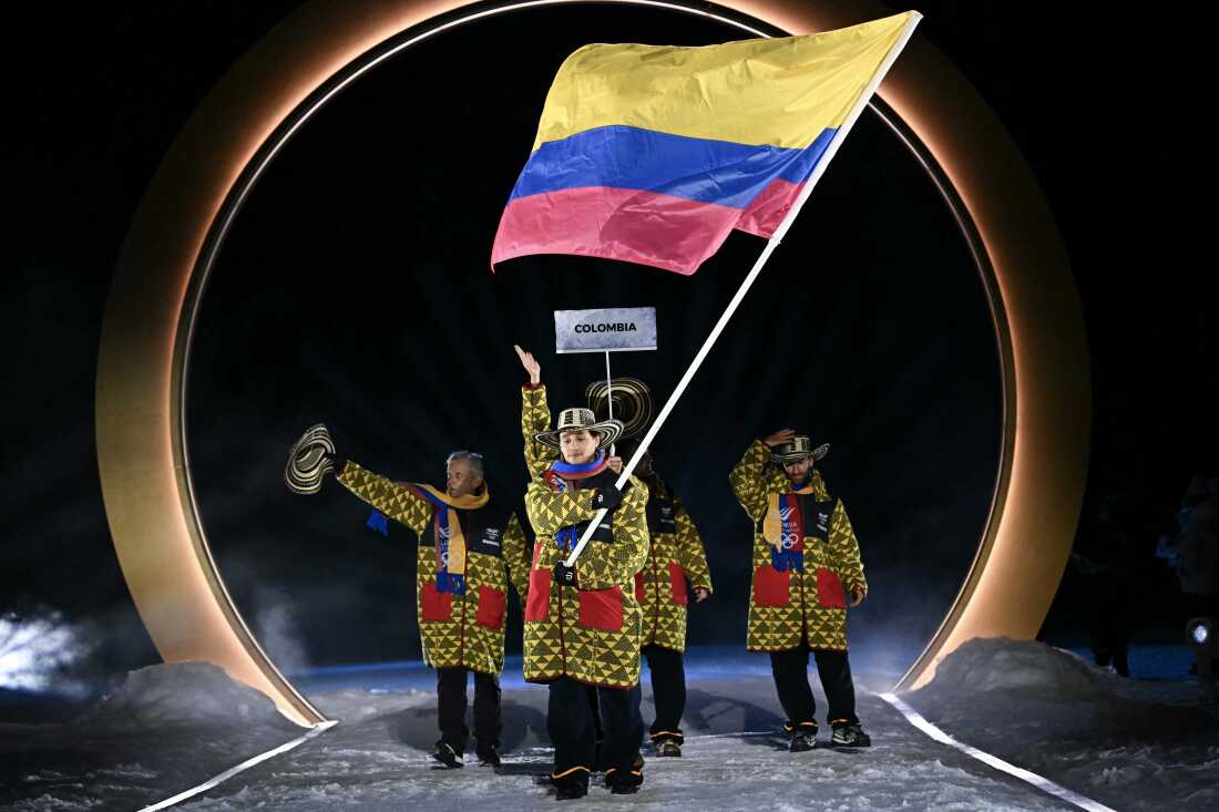 Colombia's flag bearer Fredrik Fodstad parades during the opening ceremony. He is wearing a winter jacket with a bright and dark green triangle pattern and a striped wide brim hat.