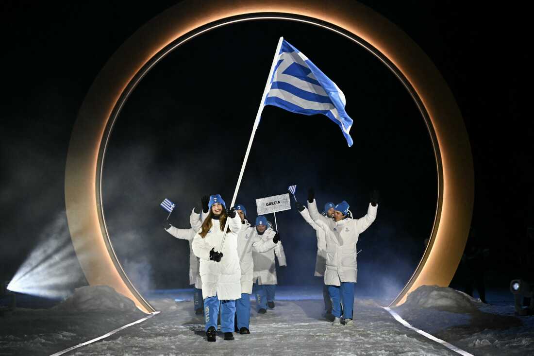 Greece's flag bearer Nefeli Tita and about 10 other athletes representing Greece parade during the opening ceremony. The athletes are wearing white winter coats and light blue pants and matching winter hats.