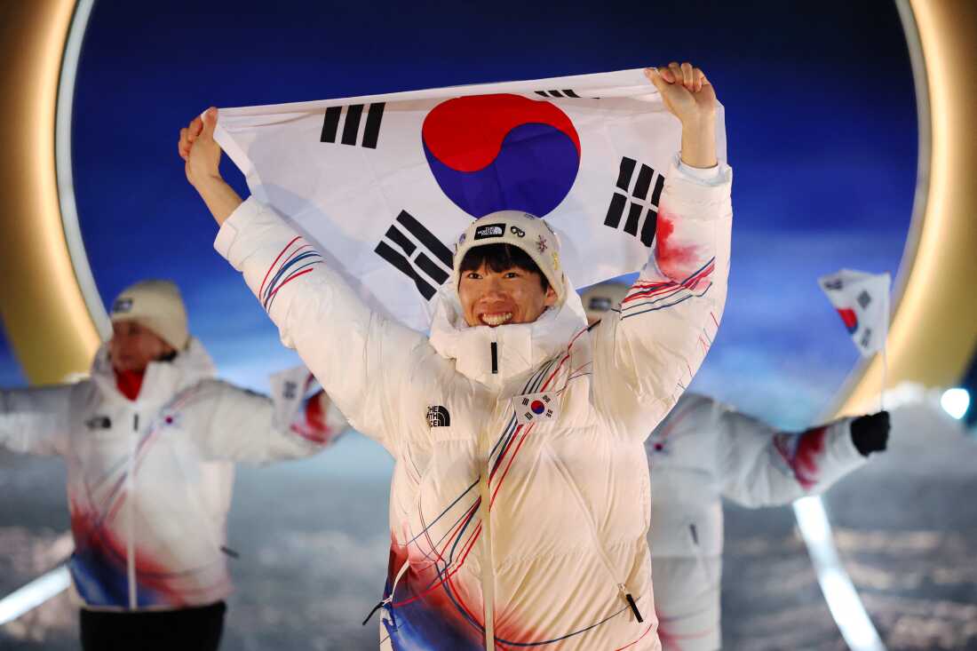 A tight photo of a South Korean athlete holding the country's flag with two fellow athletes behind. They are wearing white winter jackets with red and blue stripe design and white winter hats.