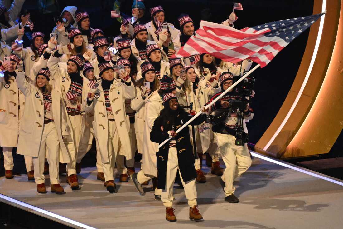 USA's flag bearer Erin Jackson parades with other athletes at the San Siro stadium. The athletes are wearing white jackets with white pants and red, white and blue knit sweaters and matching hats.